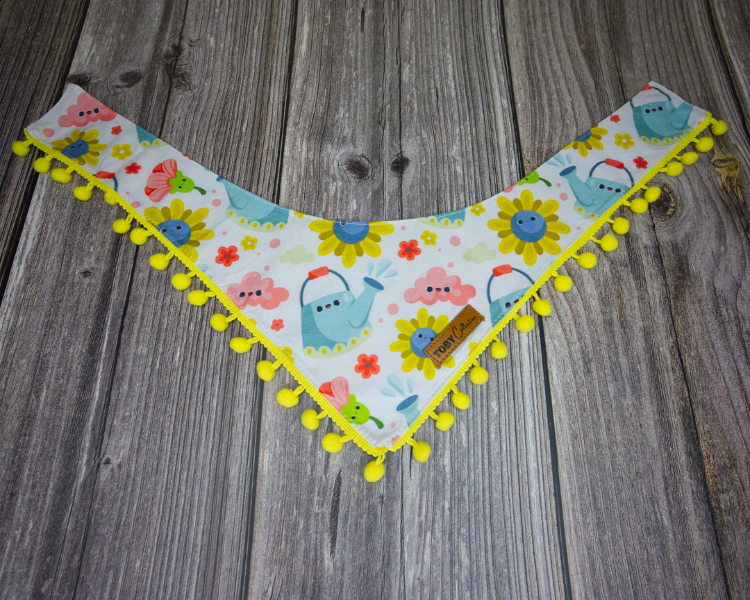 Yellow pom-pom dog bandana with a bluebird and cloud pattern on a wooden background.