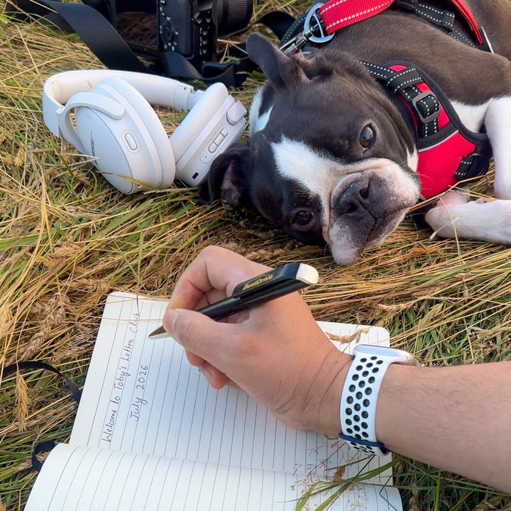 A Boston terrier dog wearing a red harness lies on dry grass next to a person writing in a notebook outdoors. A camera, white headphones, and the dog are visible in the scene.